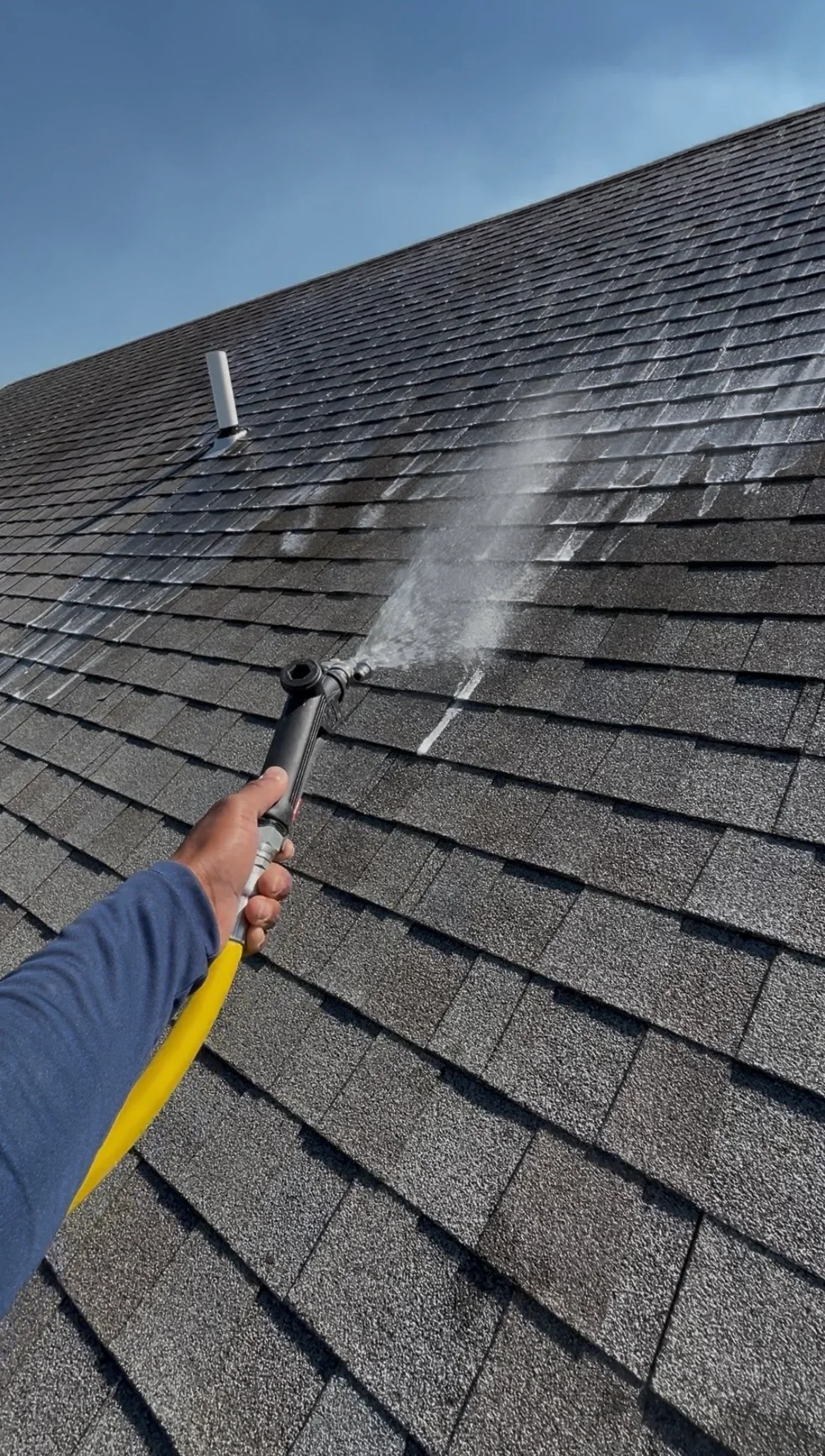 Technician soft washing dark asphalt shingle roof with low-pressure spray equipment