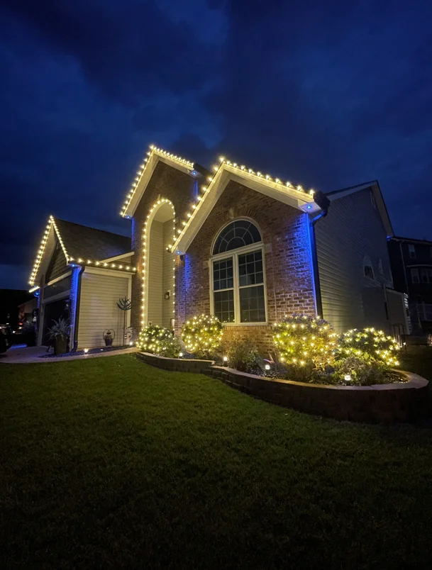 Brick home with warm LED roofline and gable lighting, blue column accents, and landscape uplighting at dusk