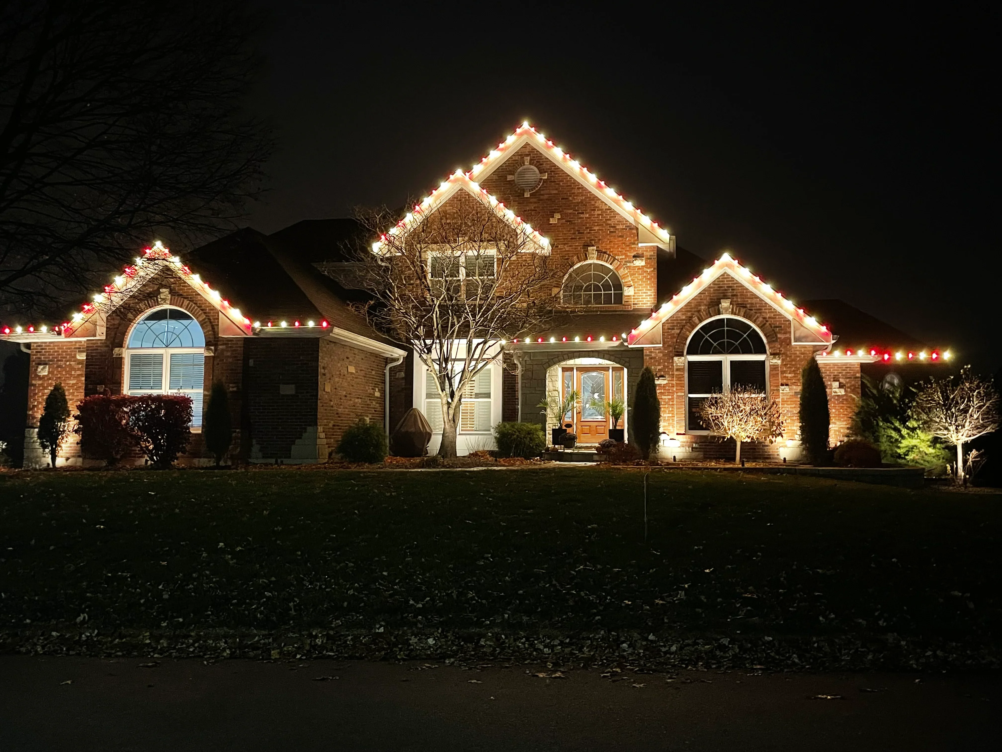 Brick home at night with warm white and red holiday lights outlining rooflines and gables