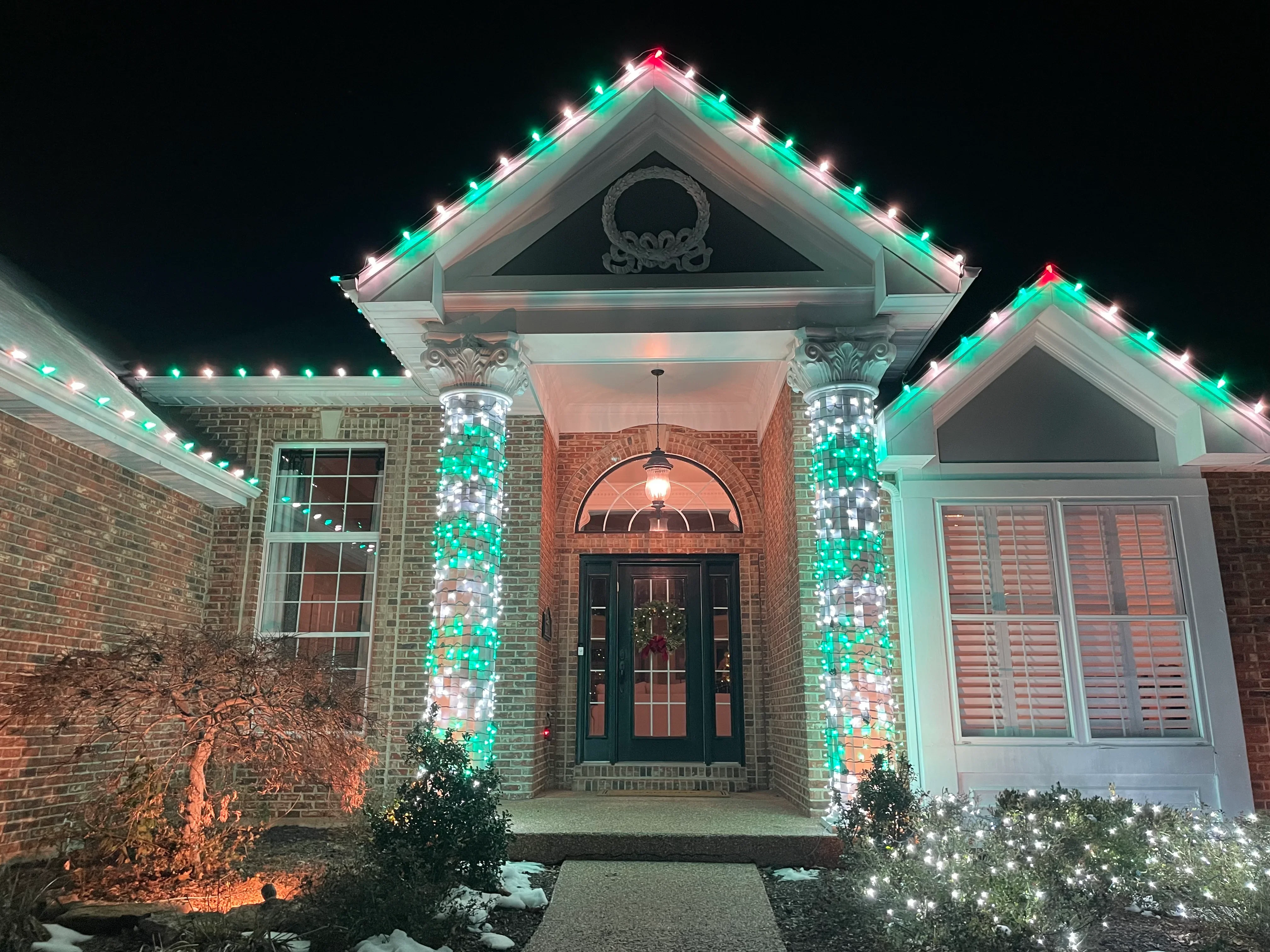 Brick home with green, white, and pink holiday lights on columns and roofline at night