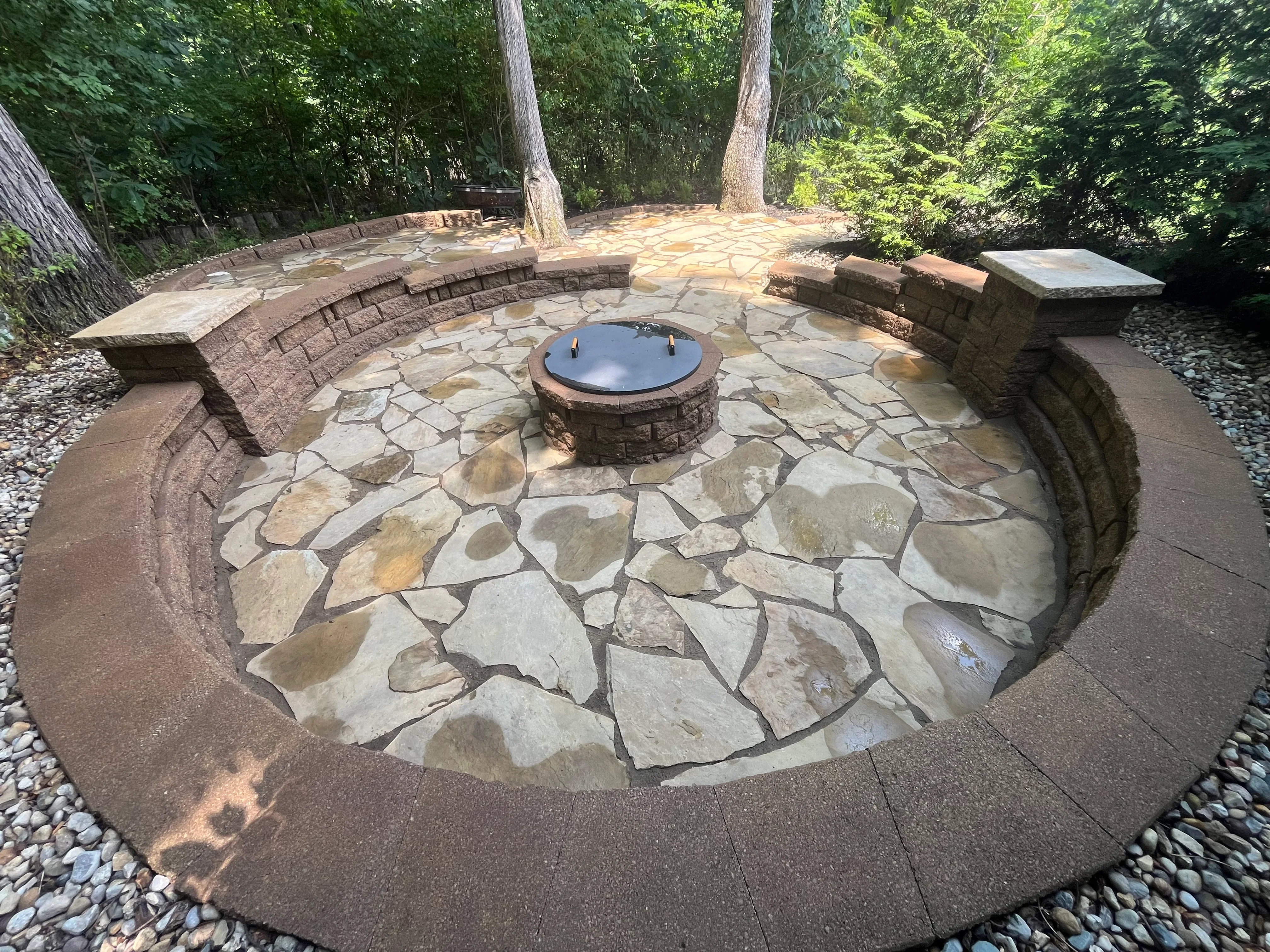 Clean flagstone patio with central fire pit and curved stone seating walls in wooded setting