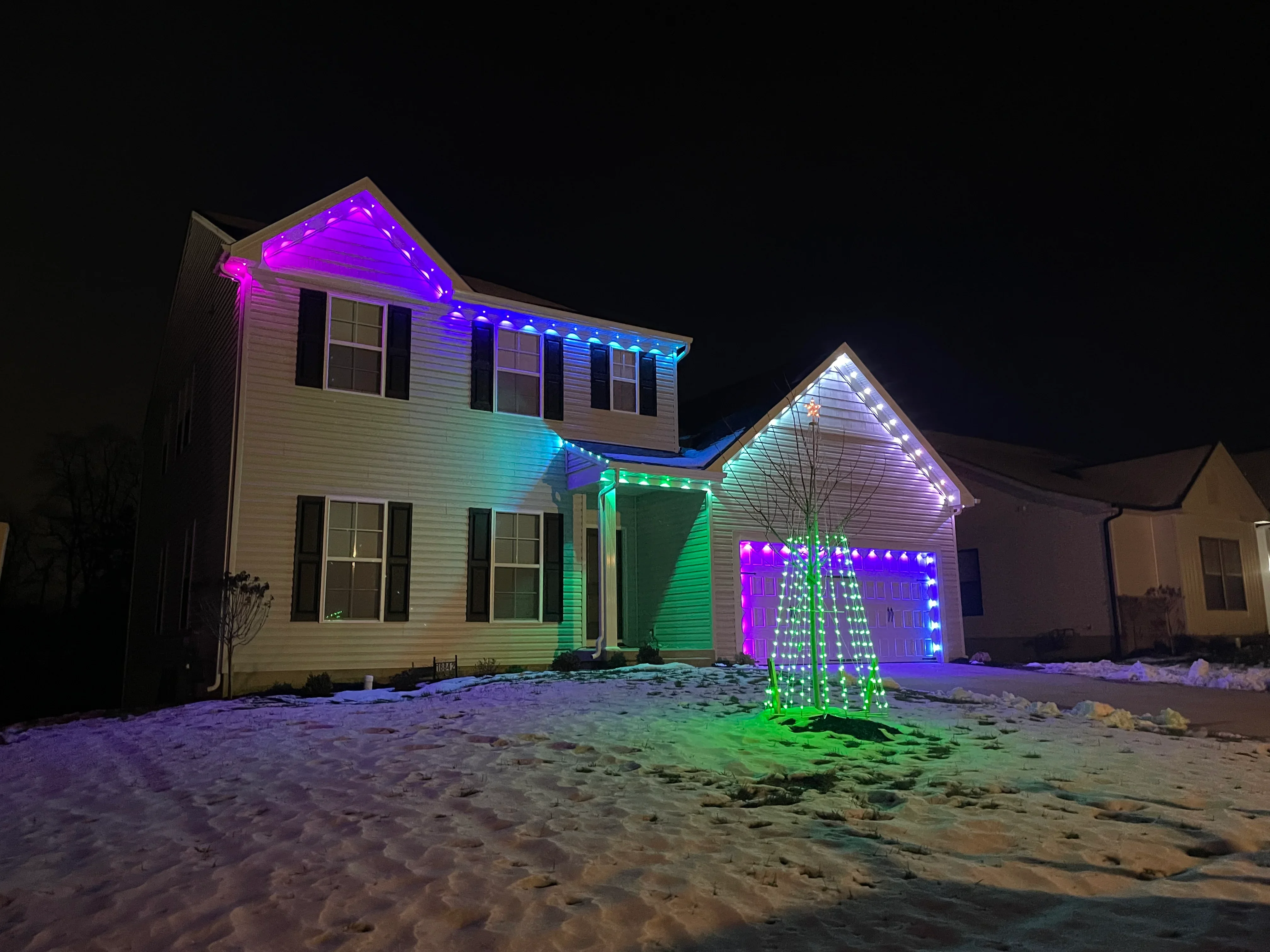 Two-story home at night with purple, blue, and green LED holiday lights on roofline and garage