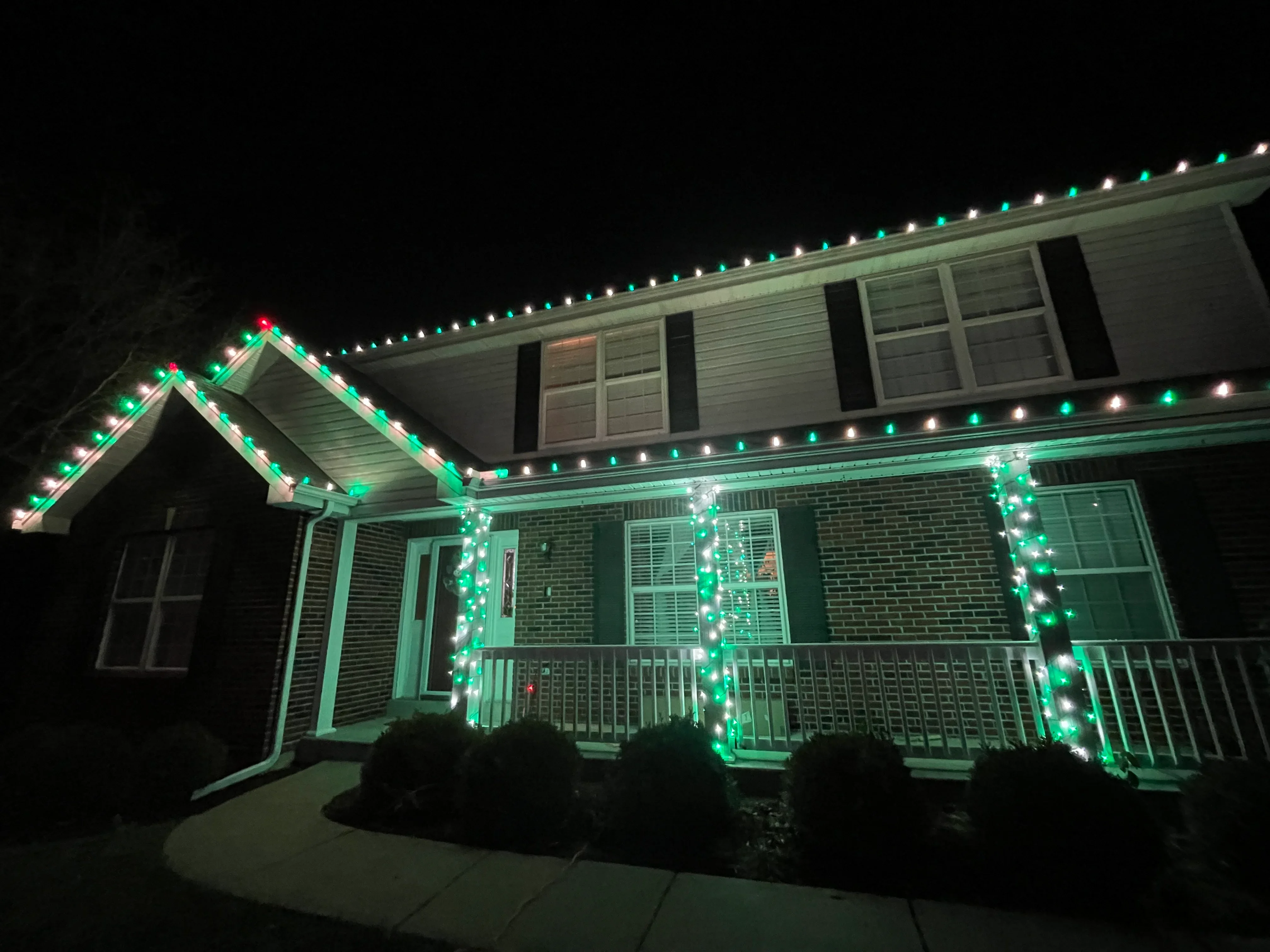 Two-story brick home decorated with green and white holiday lights along roofline and porch