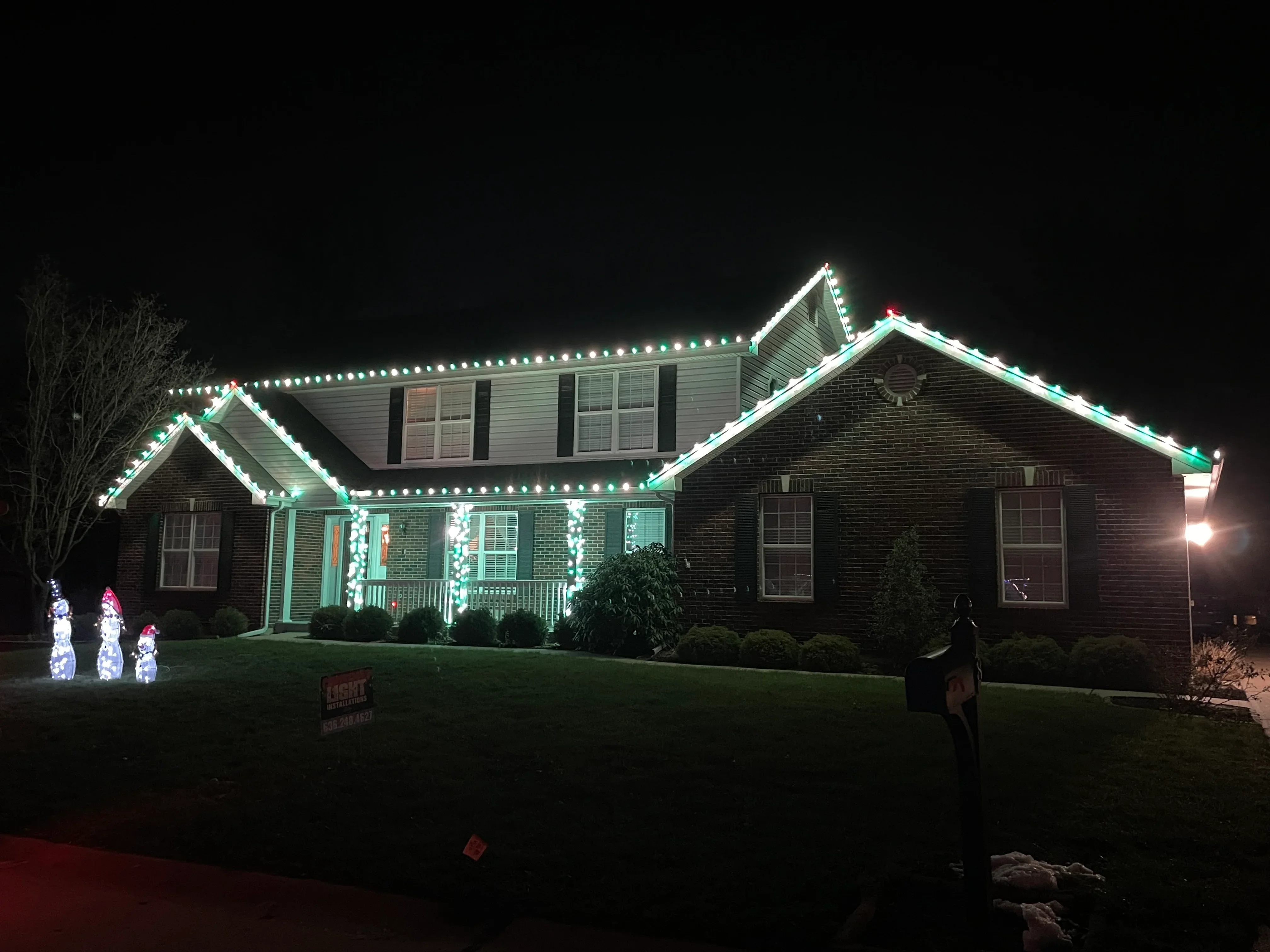 Two-story brick home with green and white LED holiday lights outlining roofline and porch at night